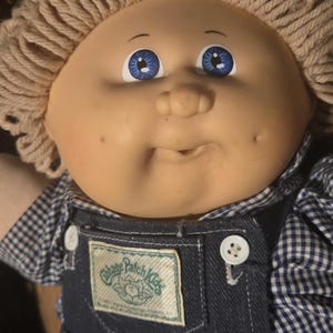 May include: A vintage Cabbage Patch Kids doll with a round face, blue eyes, and blonde yarn hair. The doll wears denim overalls with a Cabbage Patch Kids patch and a blue and white checkered shirt.