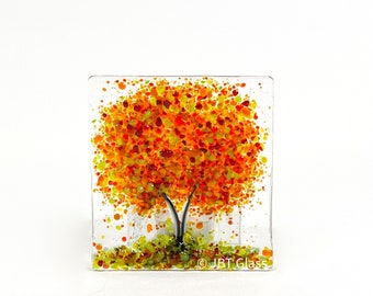 May include: A square glass art piece featuring a stylized tree with orange, yellow, and green glass leaves. The tree is set against a white background. The text "JBT Glass" is visible in the bottom right corner.