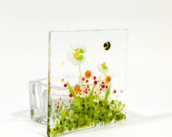 May include: A clear glass votive candle holder with a fused glass panel featuring a garden scene. The panel has white flowers, orange accents, red berries, green foliage, and a yellow and black bee. The text "© JBT Glass" is visible.