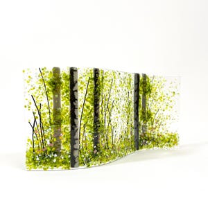 May include: A curved glass artwork showing a forest scene. The piece features black tree trunks with green foliage and small pink and white flowers. The glass is clear with a wavy design.