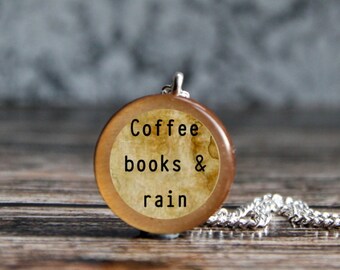 May include: A circular wooden pendant with the text "Coffee books & rain" on a cream background. It hangs from a silver chain. The pendant rests on a wooden surface.