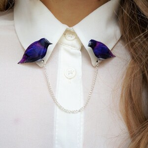 May include: A white collared shirt with a silver chain and two purple bird brooches attached to the collar.