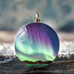 May include: A silver necklace with a round pendant featuring a vibrant image of the aurora borealis, a green and purple display of light in the night sky, with a dark silhouette of a mountain range in the foreground.
