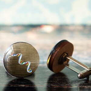 May include: A pair of cufflinks with a silver metal connector. One cufflink features a wood base with a gold-toned top and a colorful DNA strand design. The other cufflink is a plain wood base.
