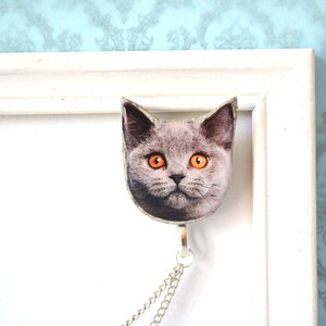 May include: A cat-shaped pendant with a silver chain. The pendant features a close-up image of a gray cat with bright orange eyes. The pendant is attached to a silver chain, and is hanging from a white picture frame.