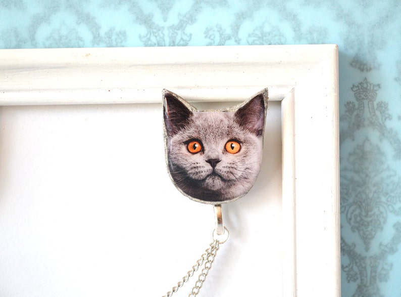 May include: A cat-shaped pendant with a silver chain. The pendant features a close-up image of a gray cat with bright orange eyes. The pendant is attached to a silver chain, and is hanging from a white picture frame.