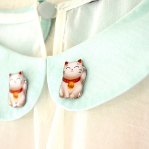 K&ouml;nnte beinhalten: Nahaufnahme einer cremefarbenen Bluse mit hellgr&uuml;nem Kragen. Zwei kleine, detaillierte Figuren einer japanischen Maneki-neko-Katze sind am Kragen befestigt. Die Katzenfiguren sind wei&szlig; mit roten und goldenen Akzenten.