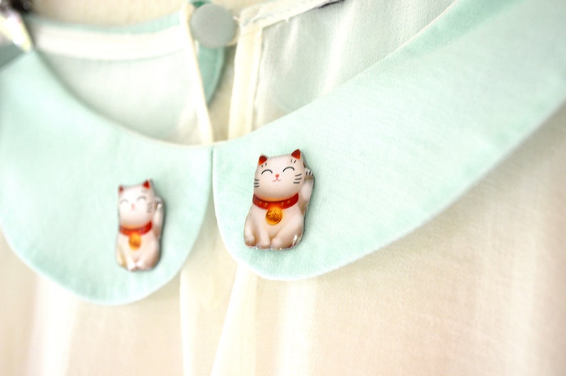 K&ouml;nnte beinhalten: Nahaufnahme einer cremefarbenen Bluse mit hellgr&uuml;nem Kragen. Zwei kleine, detaillierte Figuren einer japanischen Maneki-neko-Katze sind am Kragen befestigt. Die Katzenfiguren sind wei&szlig; mit roten und goldenen Akzenten.