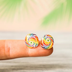 May include: A pair of colorful lollipop earrings with a rainbow swirl design. The earrings are small and round, and they are shown on a finger.