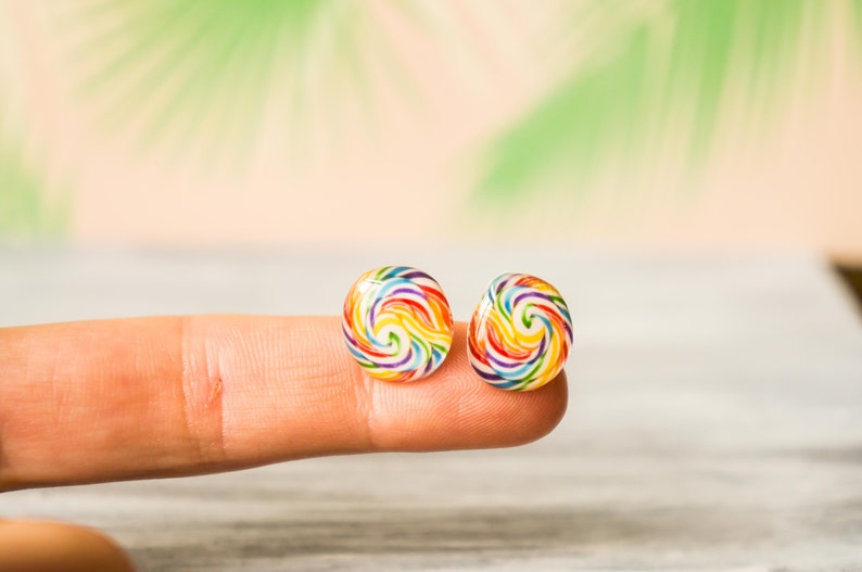 May include: A pair of colorful lollipop earrings with a rainbow swirl design. The earrings are small and round, and they are shown on a finger.