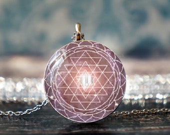 May include: A silver chain necklace with a round pendant featuring a pink and white Sri Yantra symbol. The pendant has a detailed design with a white mandala and a white Sri Yantra in the center.
