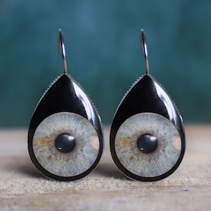 May include: A pair of teardrop-shaped earrings with a black background and a realistic eye image in the center. The eyes are a light gray color with a dark brown iris and a black pupil.