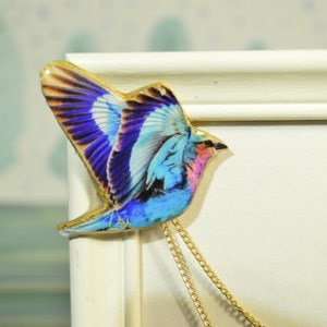 May include: A colorful bird-shaped pendant with a gold-colored trim and a gold chain. The bird features blue, purple, and pink hues in its wings and body. The pendant is likely made of a glossy material.