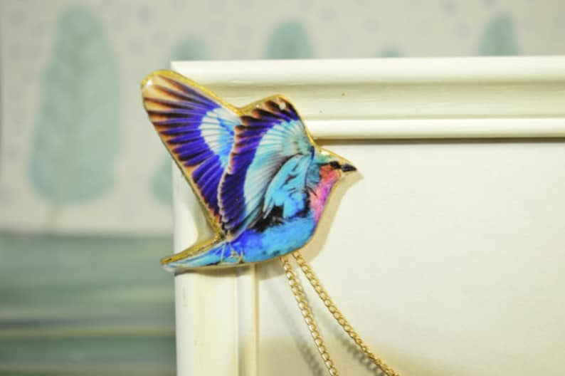 May include: A colorful bird-shaped pendant with a gold-colored trim and a gold chain. The bird features blue, purple, and pink hues in its wings and body. The pendant is likely made of a glossy material.