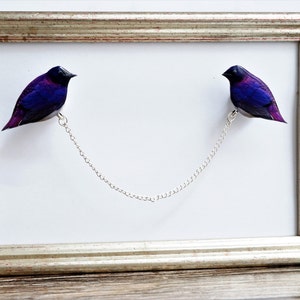 May include: Two purple and black birds are connected by a silver chain, hanging in a silver frame. The birds are facing each other.