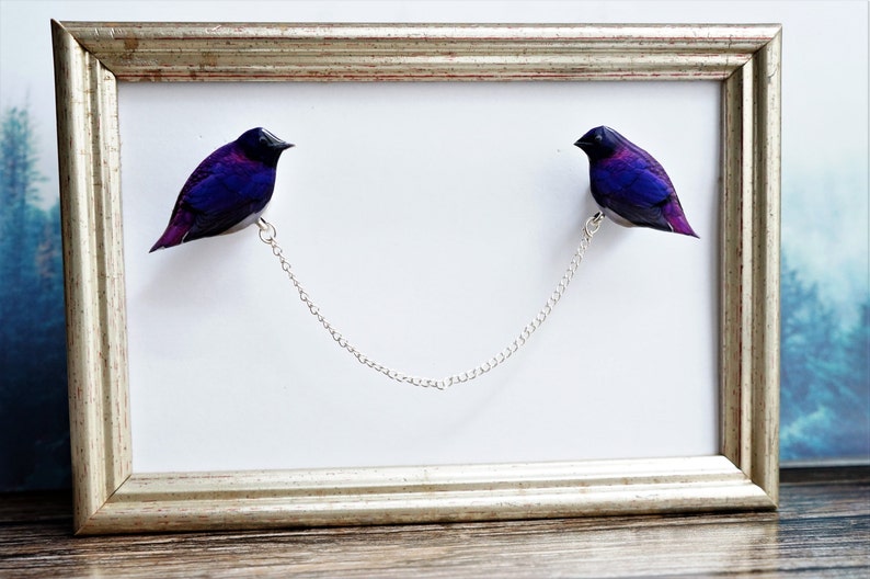 May include: Two purple and black birds are connected by a silver chain, hanging in a silver frame. The birds are facing each other.