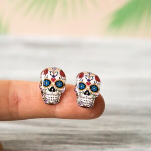May include: A pair of sugar skull earrings with white, red, blue, and black details. The earrings are made of glass and have a shiny finish.