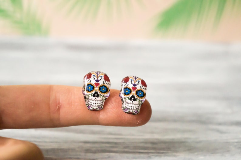May include: A pair of sugar skull earrings with white, red, blue, and black details. The earrings are made of glass and have a shiny finish.