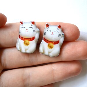 K&ouml;nnte beinhalten: Zwei kleine, wei&szlig;e Maneki Neko-Figuren mit erhobenen Pfoten. Jede Katze hat ein rotes Halsband mit einer Goldm&uuml;nze, schwarze Schnurrhaare und ein l&auml;chelndes Gesicht. Die Figuren sind etwa 2,5 cm hoch.