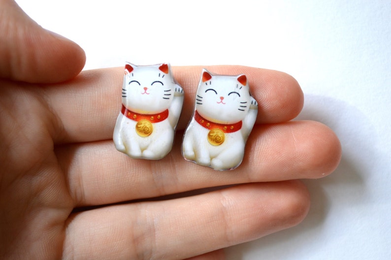 K&ouml;nnte beinhalten: Zwei kleine, wei&szlig;e Maneki Neko-Figuren mit erhobenen Pfoten. Jede Katze hat ein rotes Halsband mit einer Goldm&uuml;nze, schwarze Schnurrhaare und ein l&auml;chelndes Gesicht. Die Figuren sind etwa 2,5 cm hoch.