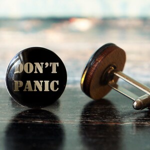 May include: A pair of cufflinks with a black, glossy face. The text "DON'T PANIC" is printed in silver. The back of the cufflink is brown and silver. The cufflinks are on a dark wooden surface.