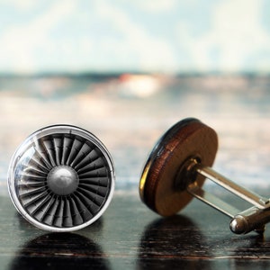 May include: A pair of cufflinks with a jet engine design. The cufflinks have a silver-tone metal base with a circular face. One face displays a detailed turbine fan, while the other has a brown wooden backing.