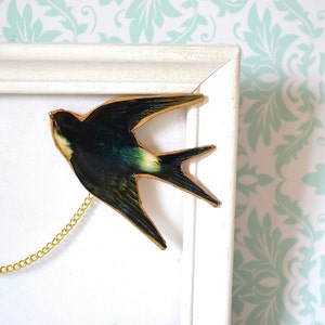 Pode incluir: Uma obra de arte emoldurada com uma andorinha com uma corrente dourada. O p&aacute;ssaro &eacute; principalmente verde escuro com detalhes em amarelo e branco. A moldura &eacute; branca e o fundo &eacute; um padr&atilde;o floral em branco e azul claro.