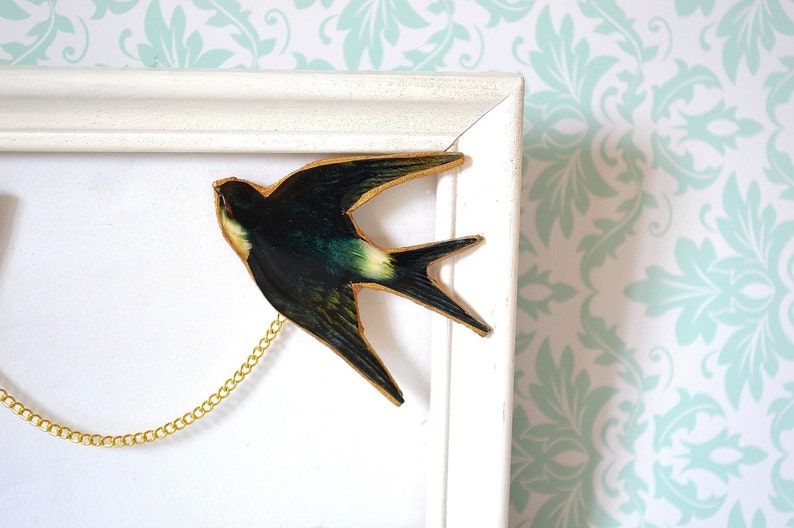 Pode incluir: Uma obra de arte emoldurada com uma andorinha com uma corrente dourada. O p&aacute;ssaro &eacute; principalmente verde escuro com detalhes em amarelo e branco. A moldura &eacute; branca e o fundo &eacute; um padr&atilde;o floral em branco e azul claro.