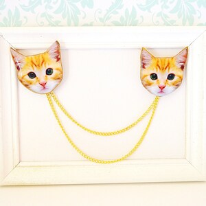 May include: Two gold chains with a cat face charm on each end. The charms are orange and white and have a realistic cat face design. The chains are hanging from a white frame.