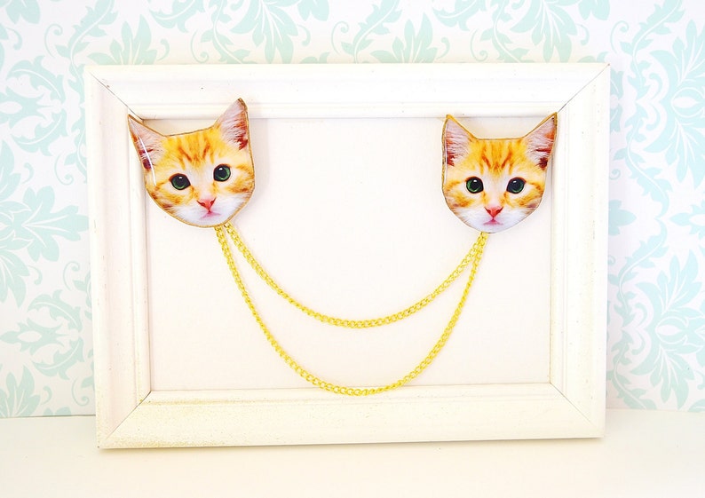May include: Two gold chains with a cat face charm on each end. The charms are orange and white and have a realistic cat face design. The chains are hanging from a white frame.