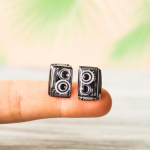 Puede incluir: Dos pendientes en miniatura con forma de cámara, negros y plateados. Cada pendiente presenta un diseño detallado con dos lentes y detalles en forma de engranaje. Son pequeños, probablemente para orejas perforadas.