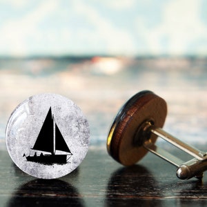 Può includere: Un paio di gemelli con un design a vela. I gemelli sono realizzati in legno e metallo. La barca a vela è in bianco e nero e ha un aspetto usurato.