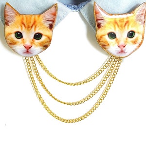 May include: A pair of gold chain necklaces with a cat face charm on each end. The cat faces are orange and white with green eyes.