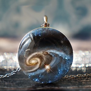 May include: A round pendant necklace featuring a galaxy design. The pendant has a clear, domed surface that magnifies the swirling colors of the galaxy within. The chain is silver-colored and delicate, complementing the celestial theme.