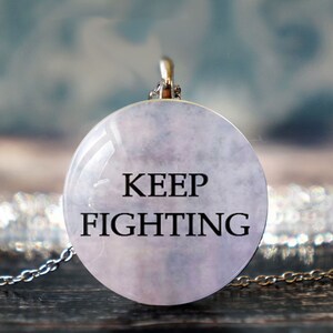 Może przedstawiać: Srebrny łańcuszek z okrągłym wisiorkiem, który ma chmurny różowy tło i napis "KEEP FIGHTING" w kolorze czarnym.