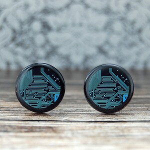 May include: Pair of black round earrings with a circuit board design in teal. The earrings are glossy and have a detailed pattern, suggesting a tech or geek theme. They are displayed on a wooden surface.