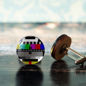 May include: A pair of cufflinks with a round, glass top featuring a black and white television test pattern. The cufflinks have a silver metal base and a brown wooden backing.