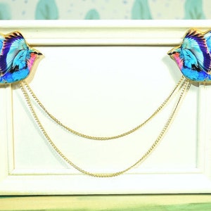 May include: A white picture frame adorned with two bird-shaped embellishments. The birds feature blue, purple, and pink wings with gold trim. Gold chains hang from each bird, creating a decorative element.