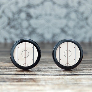 May include: A pair of black cufflinks with a white hockey rink design. The rink has a red center line and black lines for the face-off circles.