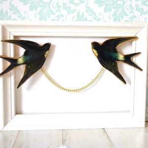Pode incluir: Uma obra de arte emoldurada com duas andorinhas conectadas por uma corrente dourada. As andorinhas s&atilde;o verde-escuras e pretas com detalhes amarelos, em um fundo branco dentro de uma moldura de madeira branca. Decora&ccedil;&atilde;o de parede.