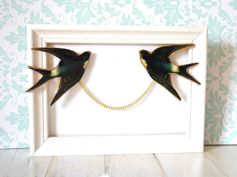 Pode incluir: Uma obra de arte emoldurada com duas andorinhas conectadas por uma corrente dourada. As andorinhas s&atilde;o verde-escuras e pretas com detalhes amarelos, em um fundo branco dentro de uma moldura de madeira branca. Decora&ccedil;&atilde;o de parede.