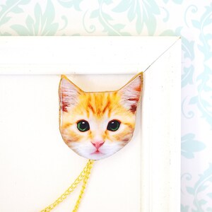 May include: A gold chain necklace with a cat face pendant. The cat has orange fur, green eyes, and a white nose.