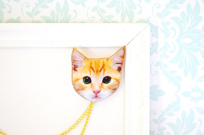 May include: A gold chain necklace with a cat face pendant. The cat has orange fur, green eyes, and a white nose.