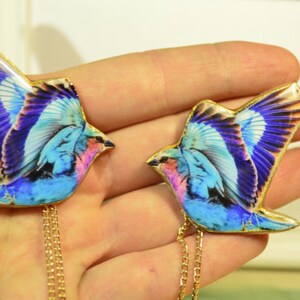 May include: Two bird-shaped pendants with gold-colored chains. The birds have blue, purple, and pink wings with gold trim. The pendants are held in a person's hand.