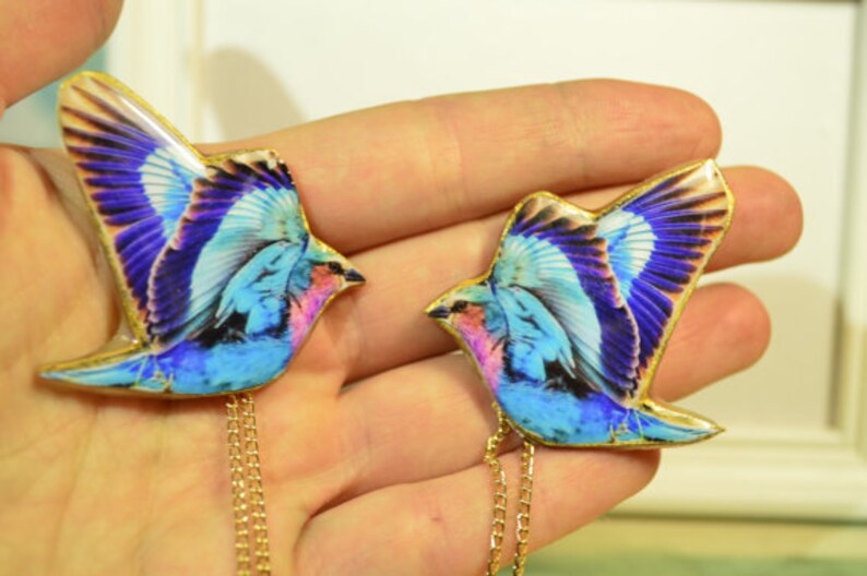 May include: Two bird-shaped pendants with gold-colored chains. The birds have blue, purple, and pink wings with gold trim. The pendants are held in a person's hand.