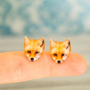 May include: A pair of fox head earrings, each featuring a realistic depiction of a fox's face in shades of orange, brown, and black. The earrings are displayed on a finger, with the fox's faces facing forward.
