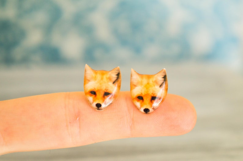 May include: A pair of fox head earrings, each featuring a realistic depiction of a fox's face in shades of orange, brown, and black. The earrings are displayed on a finger, with the fox's faces facing forward.