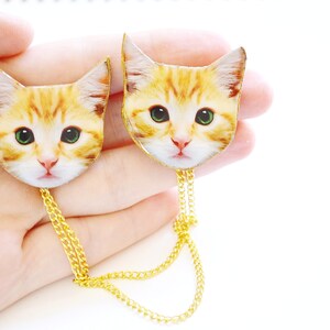 May include: Two gold chain necklaces with a cat head charm on each end. The cat heads are printed with a realistic image of a ginger cat.