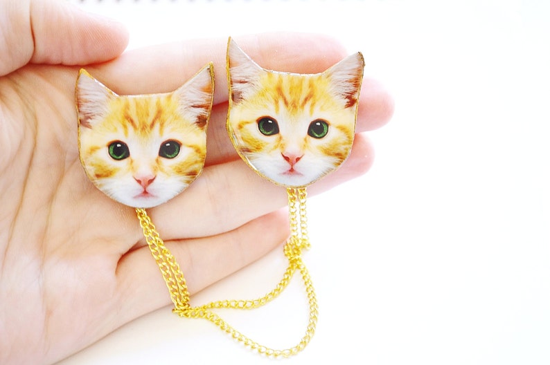 May include: Two gold chain necklaces with a cat head charm on each end. The cat heads are printed with a realistic image of a ginger cat.