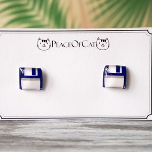 May include: A pair of square stud earrings with a retro computer diskette design. The earrings feature a white base with a blue and silver stripe detail. The earrings are displayed on a white card with the text "Peace of Cat" and a cat logo.